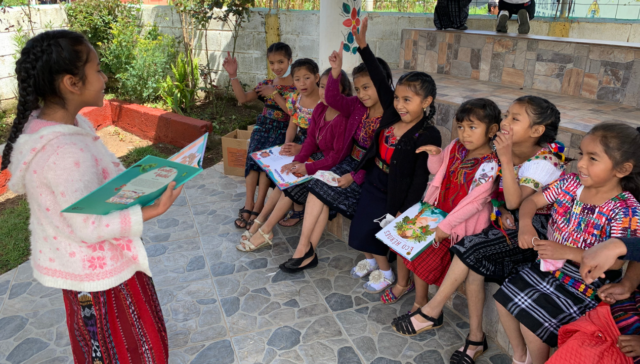 A picture of group of young  Mayan school girls with books. They are engaging with their teacher, and a few have enthusiastically raised their hands. They were brightly colored traditional clothing and are sitting outside on benches.